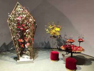 Floral coffin like display standing upright with red table and chairs next to it 